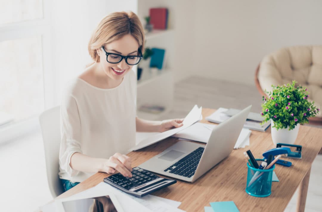 Femme souriante, travaillant sur des documents avec un ordinateur portable et une calculatrice sur un bureau, illustrant une activité de comptabilité ou de gestion administrative
