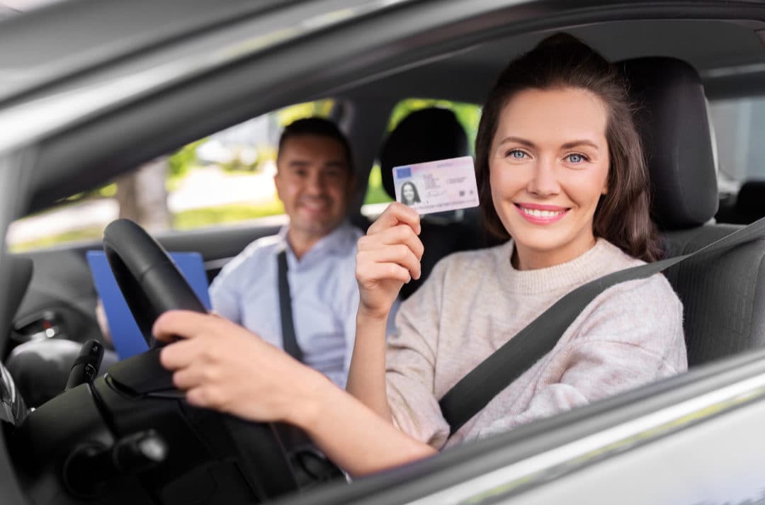 Jeune femme souriante, assise dans une voiture, montrant son permis de conduire, avec un instructeur assis à côté d'elle, illustrant l'obtention du permis de conduire.